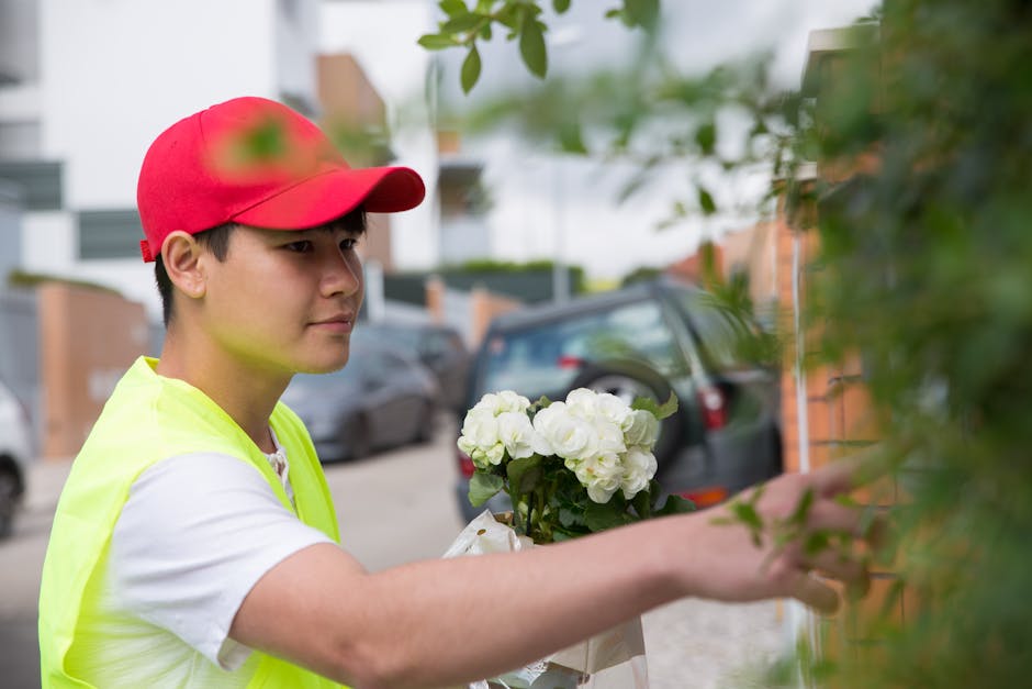 A woman with curly blonde hair, dressed in a light patterned blouse, is smiling as she receives a tall bouquet of pink roses with green foliage from a florist with her hair tied back, who is wearing a