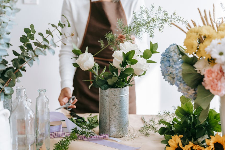 A florist, wearing a brown apron over a white long-sleeve top, arranging a bouquet of white roses with glossy green leaves and sprigs of light green foliage in a tall, rustic, galvanized metal vase on