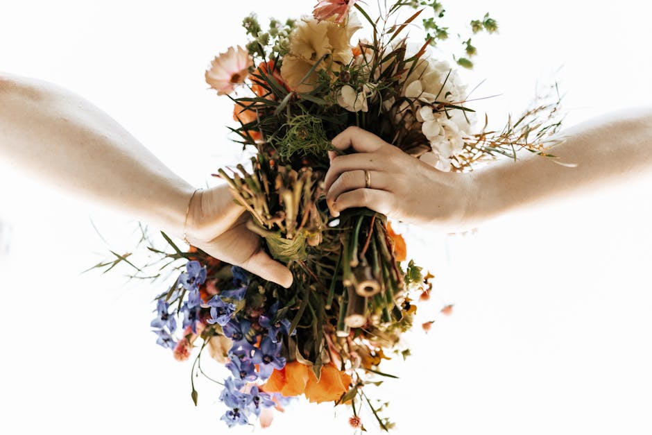 A florist, wearing a brown apron over a white long-sleeve top, arranging a bouquet of white roses with glossy green leaves and sprigs of light green foliage in a tall, rustic, galvanized metal vase on