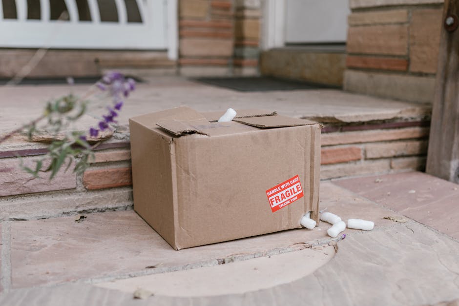 An open brown cardboard box with flaps folded outward, lined with transparent bubble wrap for protection, sitting on a plain white surface. A white and red 'FRAGILE' tape is partially unrolled near th
