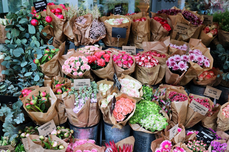 A person wearing a light grey cardigan and dark trousers is selecting a floral arrangement at a flower market. Their right hand gently touches a bouquet wrapped in clear plastic, containing purple hya