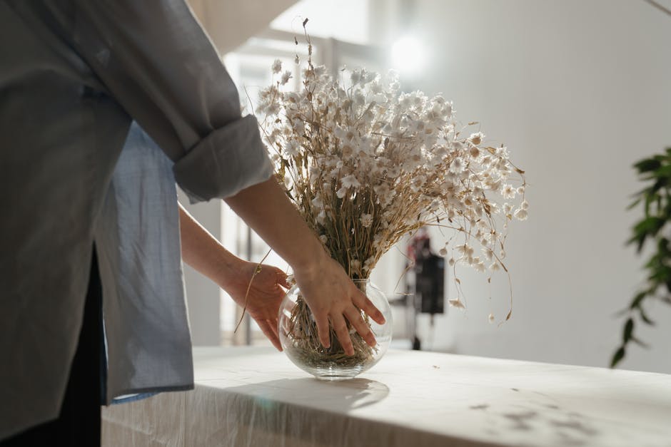 A pair of hands arranging a bouquet of fresh flowers in a clear glass jar filled with water, placed on a white surface in a well-lit workspace. The bouquet includes white and green flowers with small
