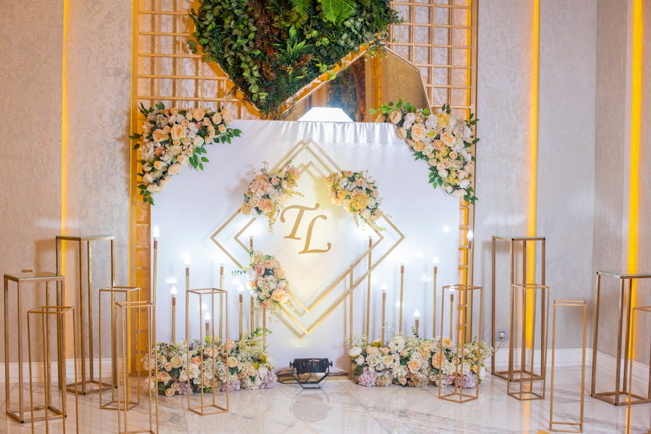 A decorative wedding backdrop featuring a white fabric background with the gold monogram initials 'TL' in the center, surrounded by pastel-colored floral arrangements of cream, blush, and peach roses,