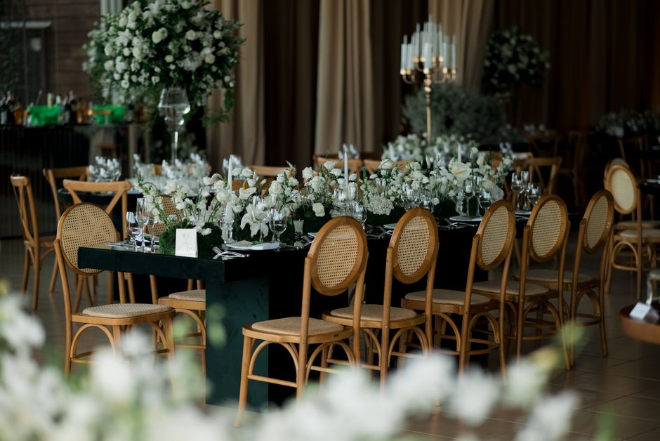 A long rectangular banquet table decorated for a wedding reception, covered with a dark tablecloth and adorned with elegant floral arrangements featuring white roses, lilies, and greenery. The flowers