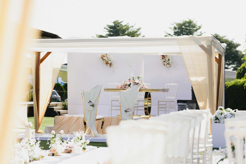 A long rectangular banquet table decorated for a wedding reception, covered with a dark tablecloth and adorned with elegant floral arrangements featuring white roses, lilies, and greenery. The flowers